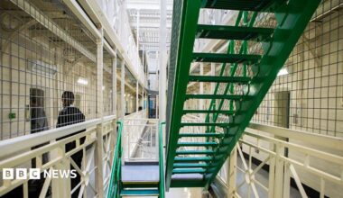 A prisoner is escorted to his cell by an officer next to a set of green stairs situated in between rows of cells on Benbow wing inside HMP/YOI Portland.