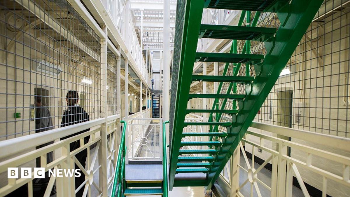 A prisoner is escorted to his cell by an officer next to a set of green stairs situated in between rows of cells on Benbow wing inside HMP/YOI Portland.