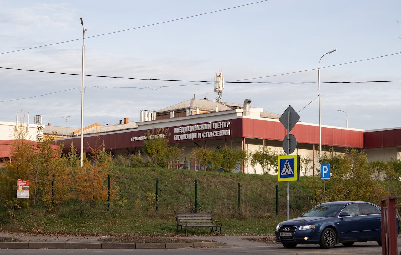 An image of a building with a white concrete building with clay red cladding and Russian words in white letters. The building sits on a grass covered hill, with a metal fence in front. A car drives along a street in front of the hill.