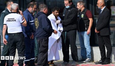 A man is wrapped in a large white blanket after being rescued from an attacked ship. There are seven people around him, one of whom has a stethoscope around his neck