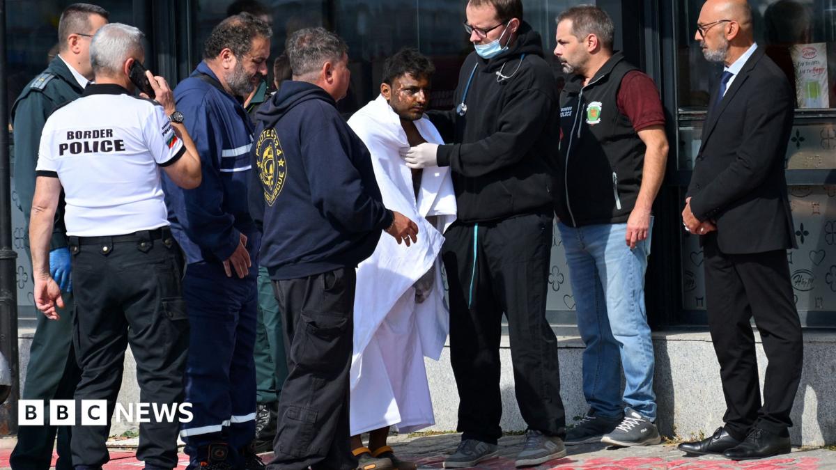 A man is wrapped in a large white blanket after being rescued from an attacked ship. There are seven people around him, one of whom has a stethoscope around his neck