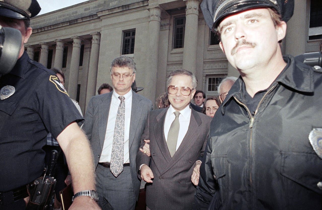 A man in a brown suit is led smiling from a building by two men. In the foreground is a police officer.