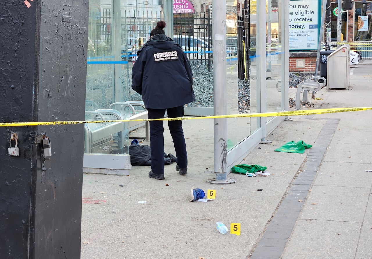 A police officer wearing a jacket with the word "forensics" on it, examines the inside of a bus shelter and places evidence tags on the ground.