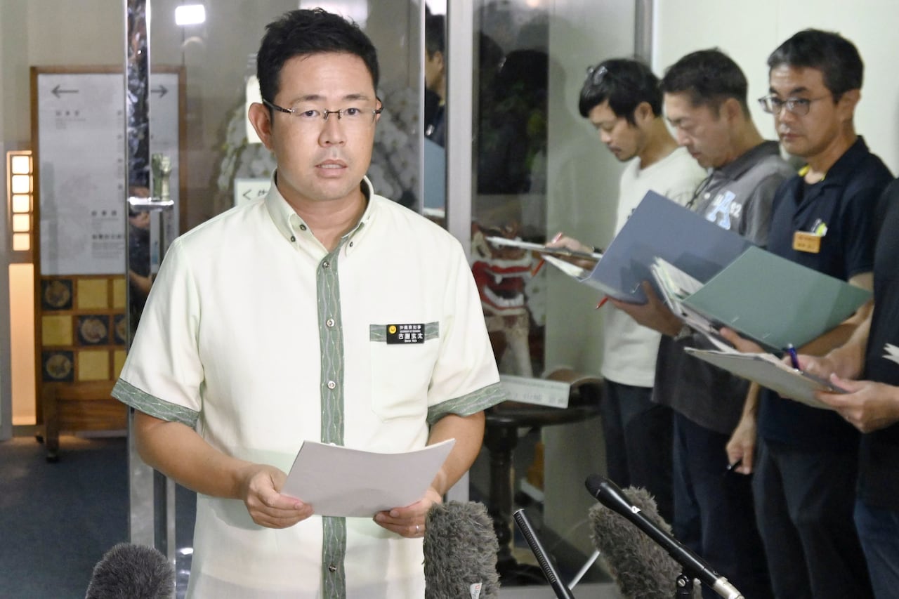 A bespectacled man in a white collared shirt reads from sheets of printed paper as he stands in front of a row of microphones. Three men stand lined up in the right corner behind him holding open folders or binders.