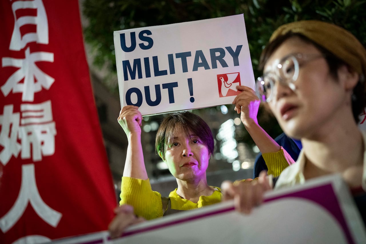 A woman stands amid protesters at night holding a printed sign above her head that reads "US Military out!"