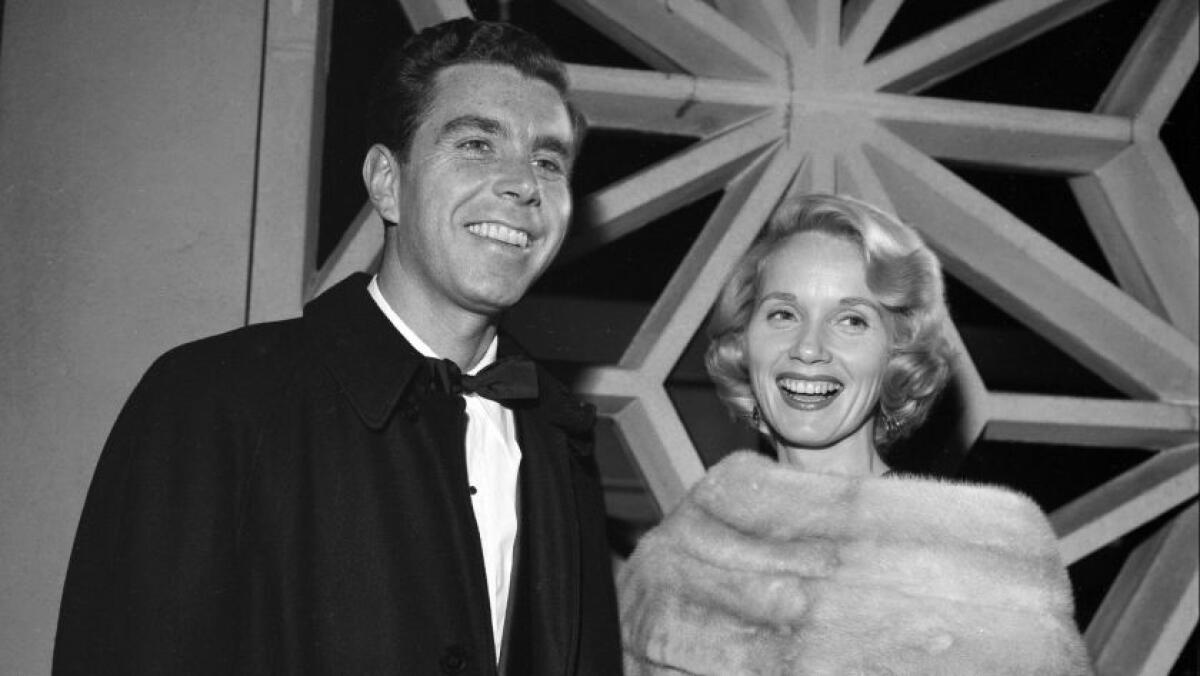 Eva Marie Saint in a fur stole and Jeffrey Hayden in a tux with a bow tie stand in front of a window.