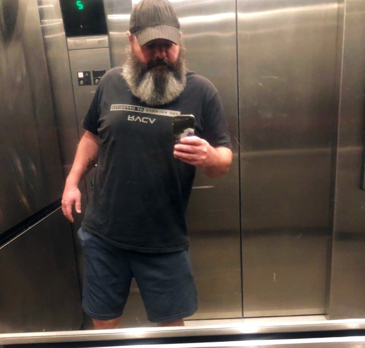 A man takes a mirror selfie in an elevator to showcase his slanted spine. He wears a ballcap, t-shirt and shorts and has a long salt and pepper beard.
