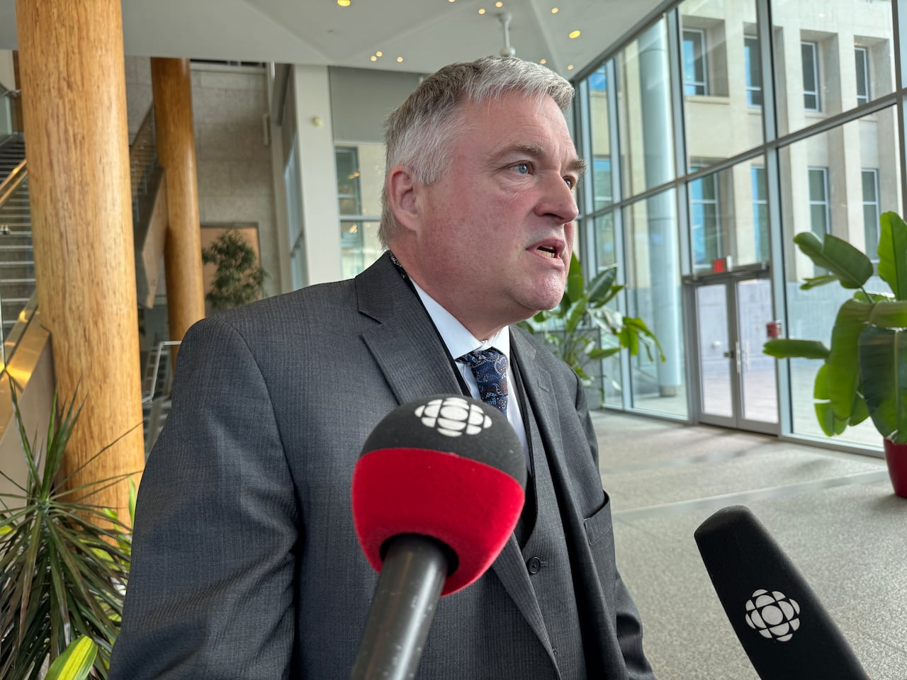 A man in a grey suit speaks with reporters with wooden columns, tall windows and plants in the background of the Queen Elizabeth II Building in Edmonton.