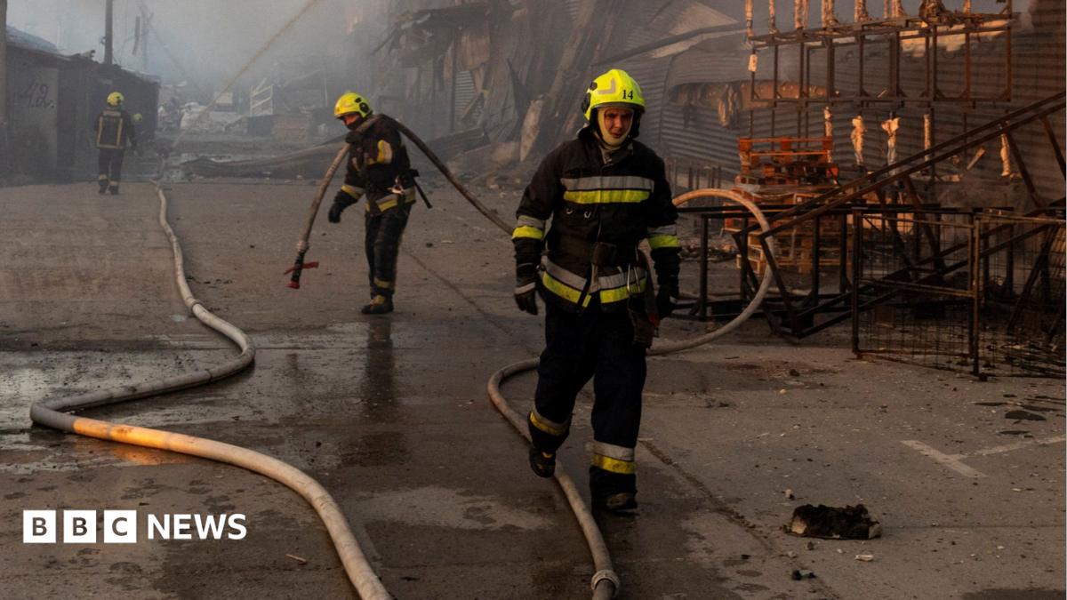 Three firefighters wearing yellow helmets work at the site of a warehouse that burned out after it was hit during a night of Russian missile attacks in Kyiv. One of the firefighters is seen holding a large hose. The destroyed remains of the warehouse can be seen in the background
