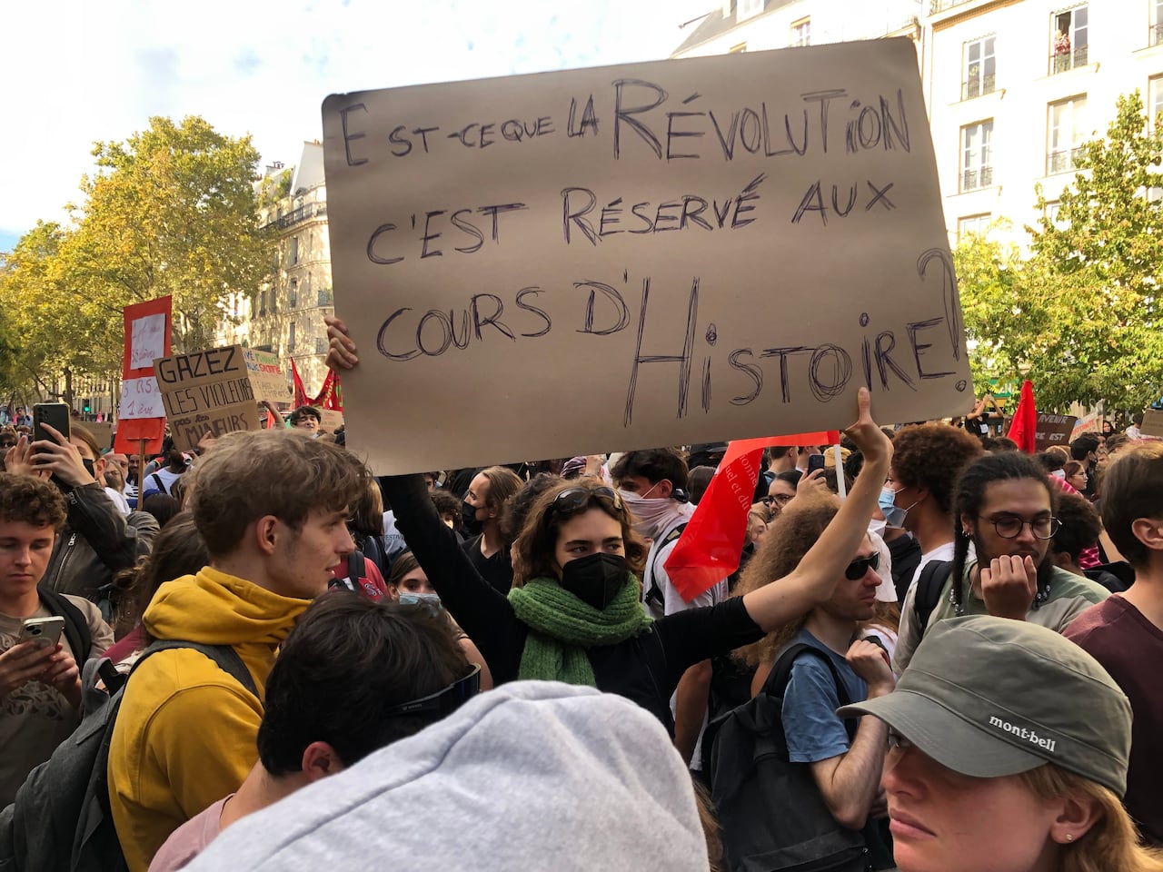 In the middle of a crowd, a protester in a black mask and green scarf holds a brown sign.