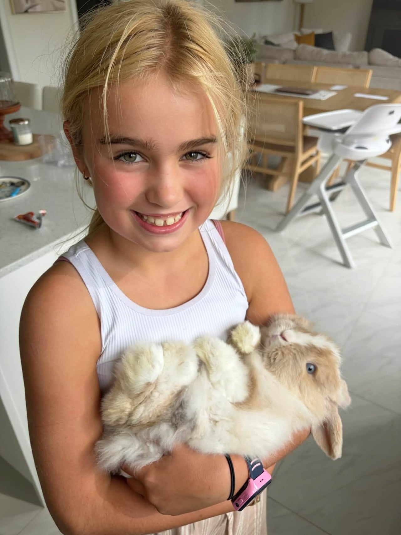 A photo of a girl with blonde hair, smiling as she holds her pet bunny, who is in her arms on his back.