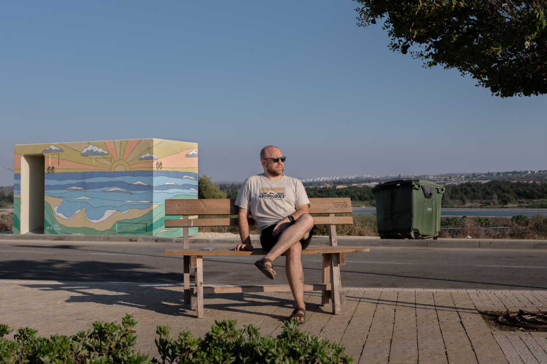 ZIKIM, ISRAEL, AUGUST 31, 2026: Shahar Goldenberg, Kibbutz Zikim's management member sits down for a portrait with a bomb shelter behind him. The city of Sderot is visible in the far distance. Credit: Amit Elkayam for NPR.