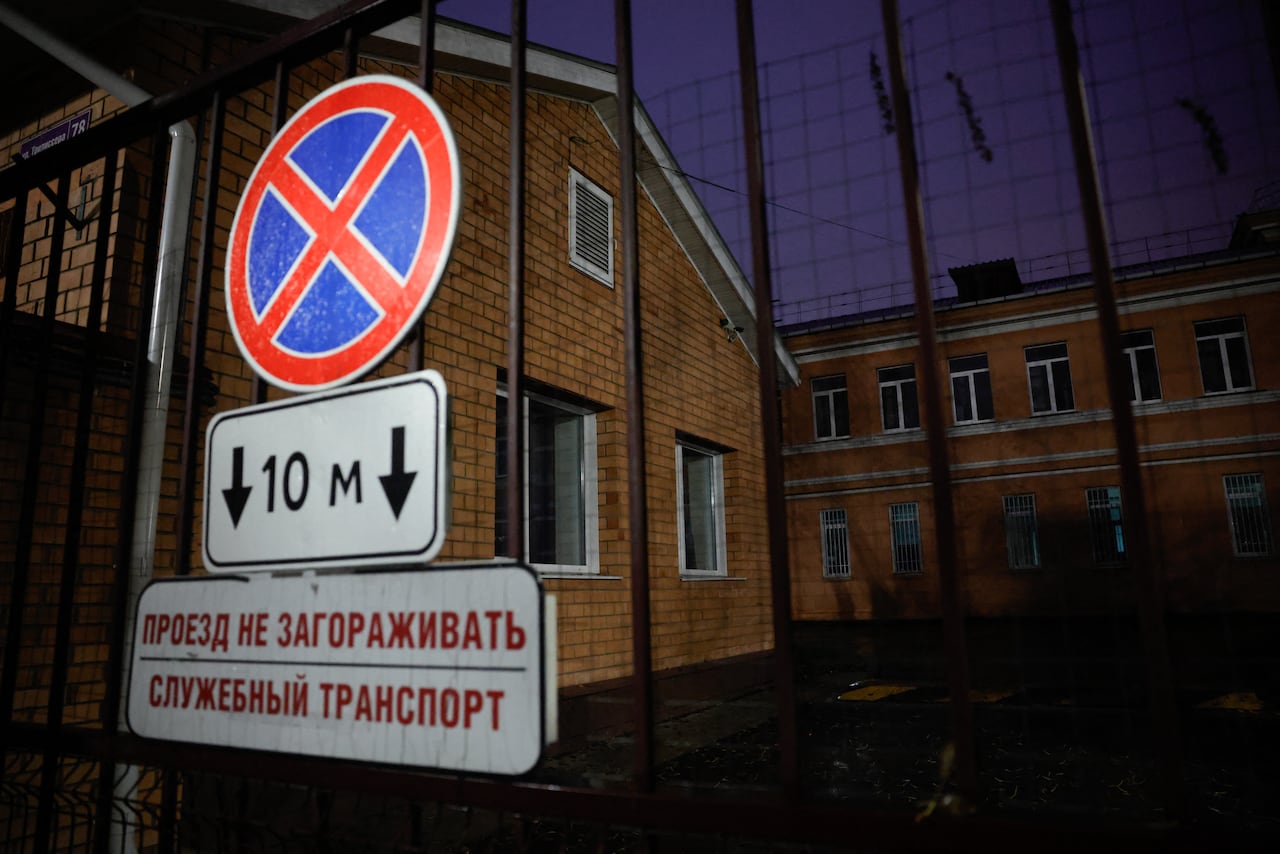 A nighttime view of a large brick building behind a metal rod gate, with a blue and red, circular sign with an X on it above another white sign with Russian words in black lettering.