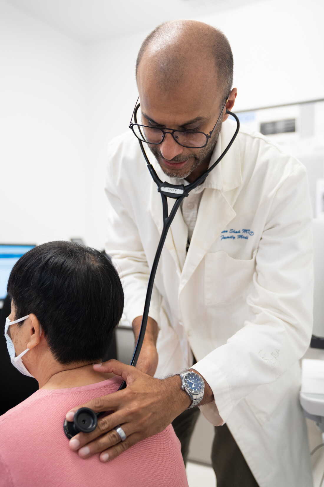 Naman Shah stands near a seated patient, holding the end of his stethoscope against her back.