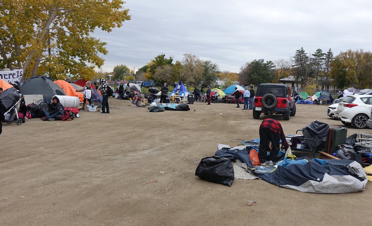Piles of belongings in a large gravel area with parked vehicles and people milling around.