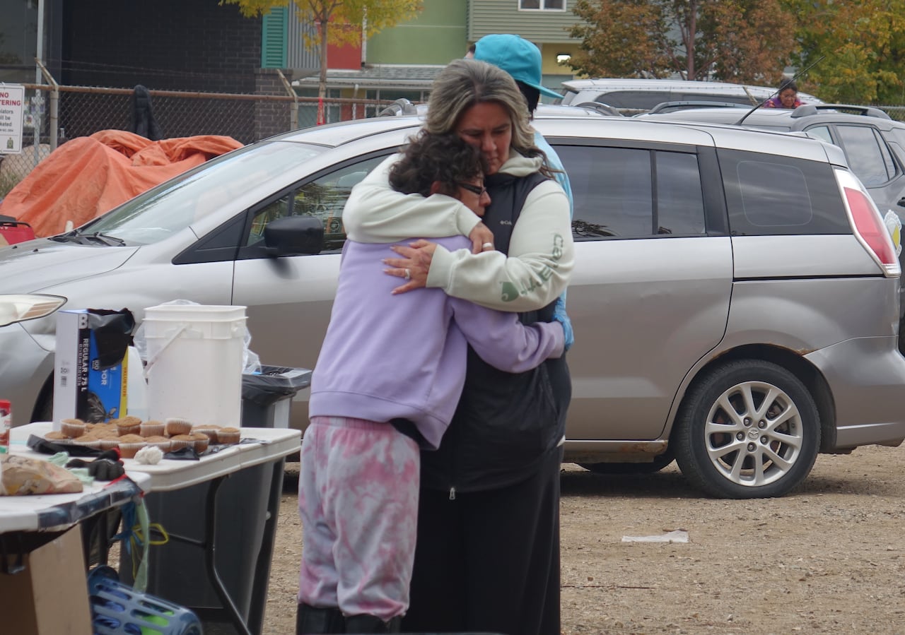 Two women embrace in a gravel parking lot next to a table of food.