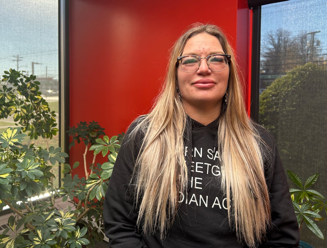 A woman wearing a black sweatshirt stands in front of a red wall, with plants in the background
