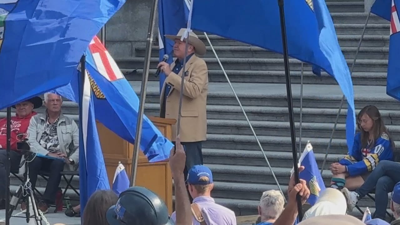 A man in a tan suit and a cowboy hat speaks into a microphone on the steps of a building. There are many Alberta flags being waved around him.