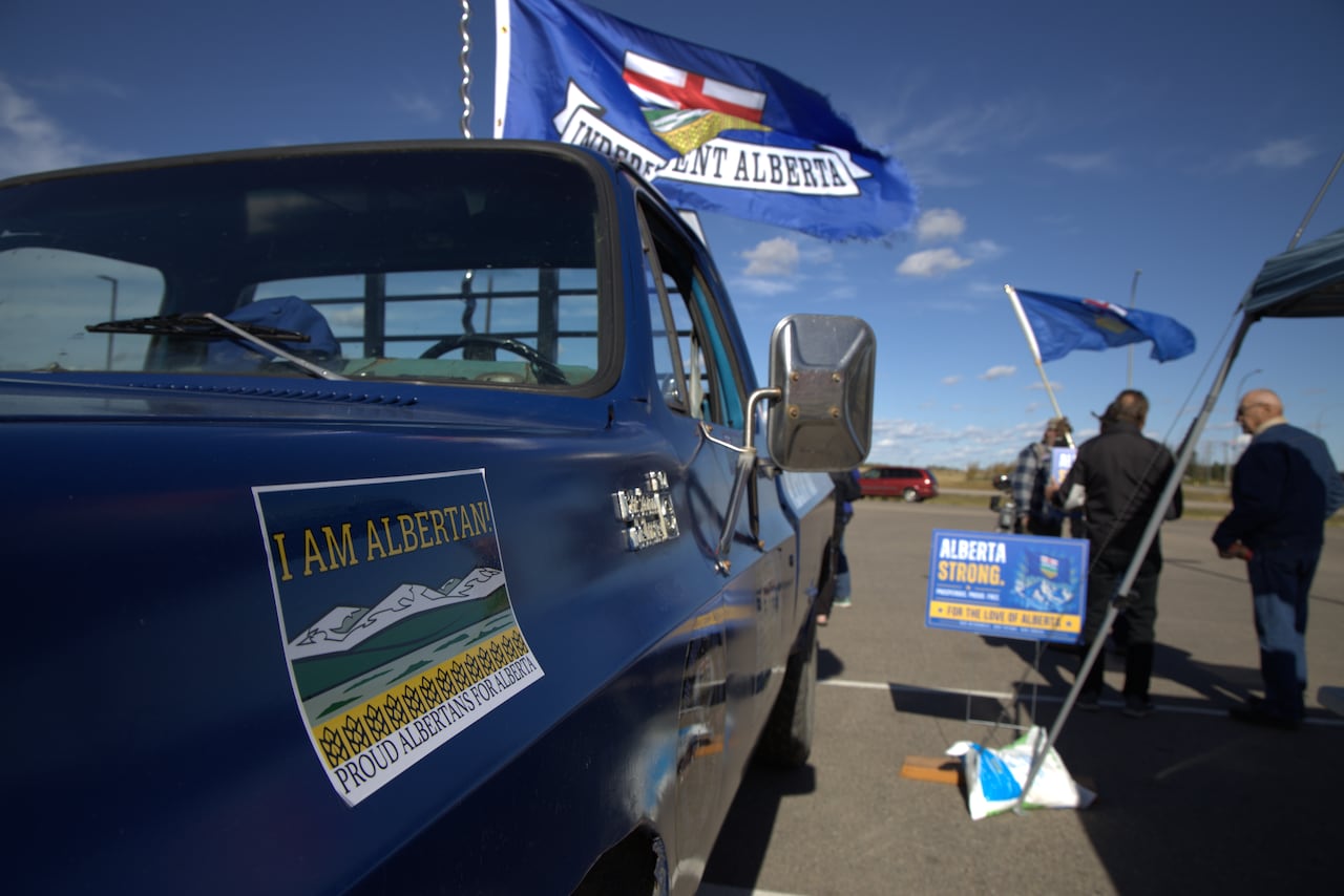 A blue truck with Alberta slogans is seen beside a small group of people. An Alberta flag and a sign are also visible.