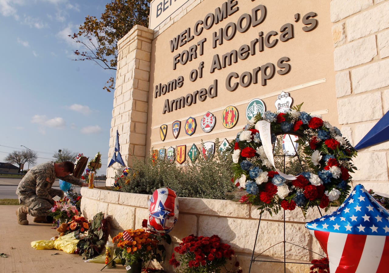 A person in military fatigues knees by a sigh that says welcome to fort hood, surrounded by flowers
