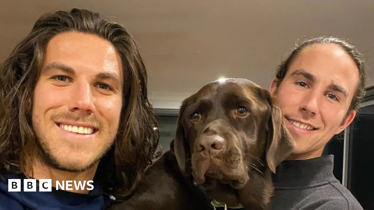 A young man with shoulder-length brown hair and light stubble smiling at the camera, and another young man to his right and a brown dog in between them