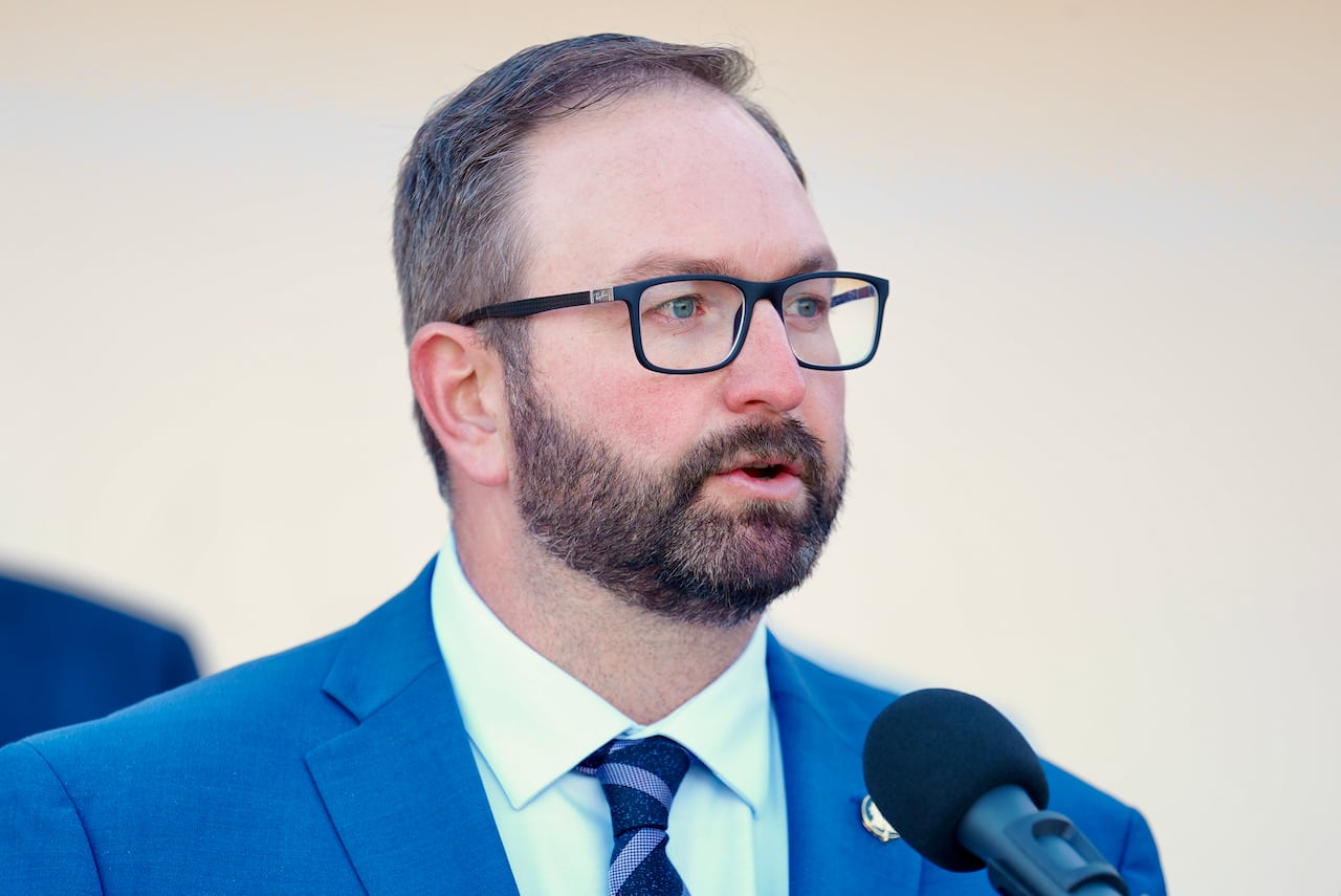 A man speaks to a microphone while in a light blue suit, white shirt and blue checkered tie.