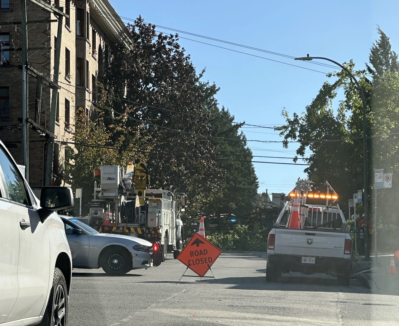 A police car and work trucks block access to a road, with foliage seen in the street behind them.