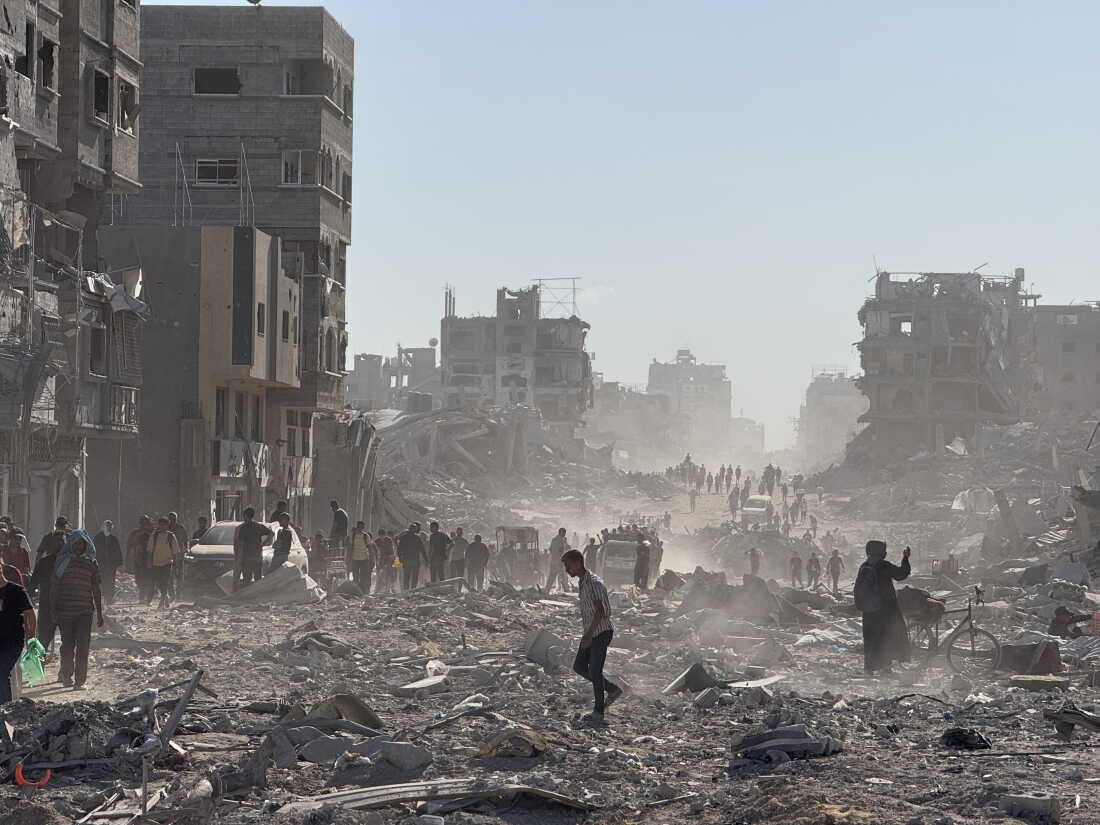 Palestinians walk amid rubble, debris and the skeletons of heavily damaged multistory buildings.