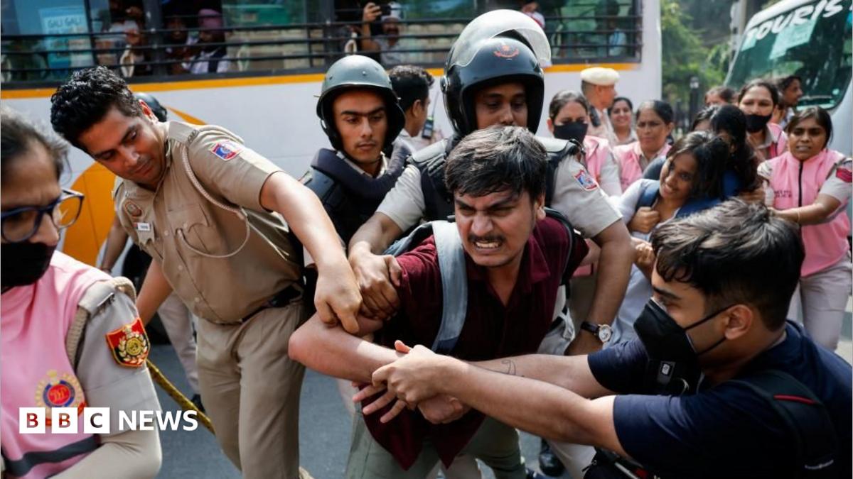 A man is being held by men in official uniforms as another man grapples with them. More people are stood behind in pink uniforms holding other protesters.