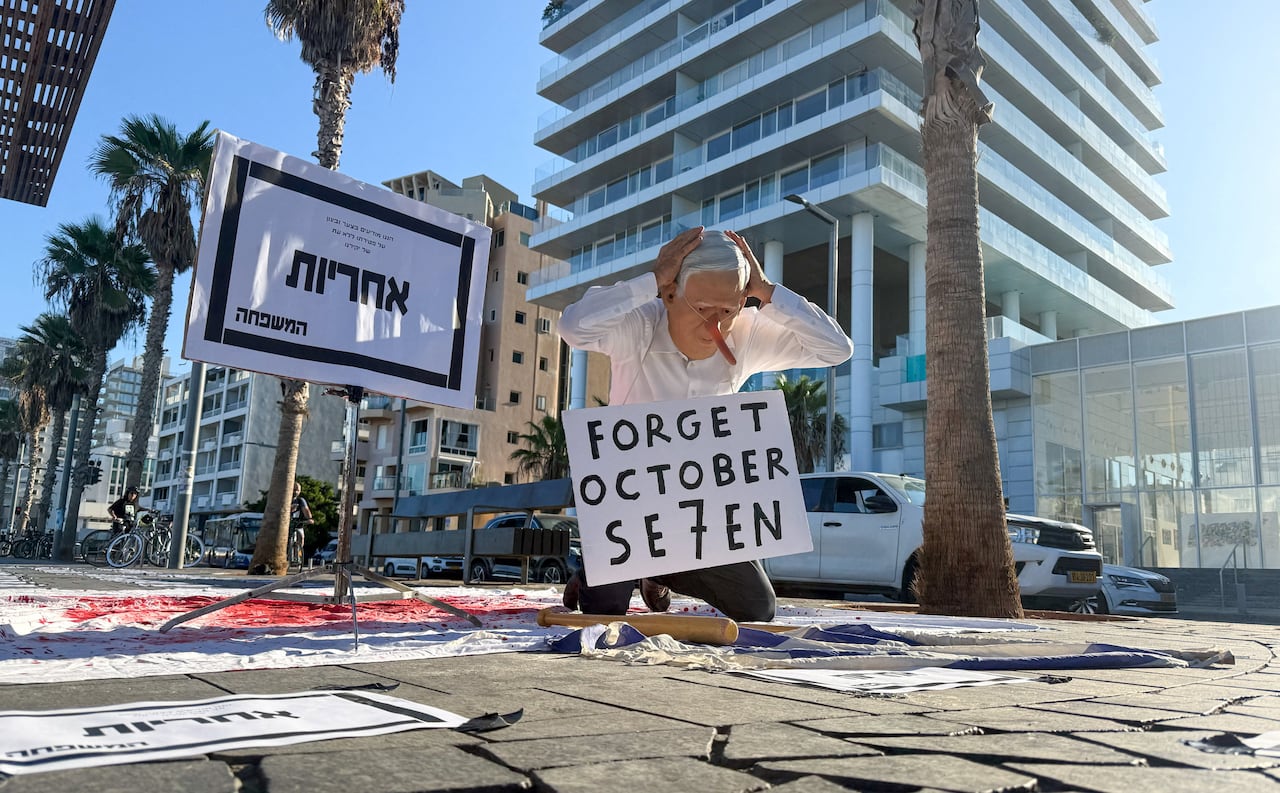 A man wearing a full-face mask with a long nose, kneels on the pavement of a city street. A placard around his neck reads 'Forget October Seven'