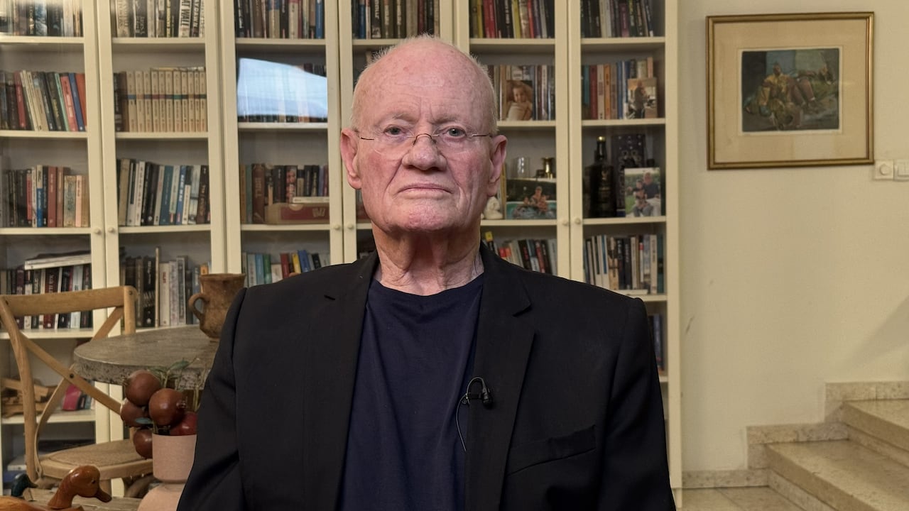 An older and stern-looking bald man poses for a photo. There's a bookcase behind him.