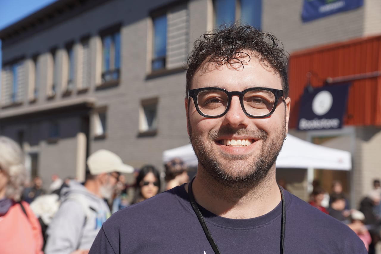 A white man with short curly black hair and glasses smiles at the camera. He's standing outside on a sunny day in front of a crowd.