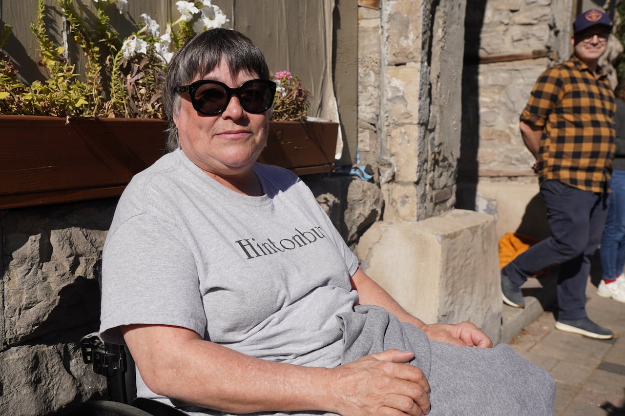 A woman in a wheelchair is seen next to a building on a sunny day. She is wearing black sunglasses and a grey shirt reading "Hintonburg."