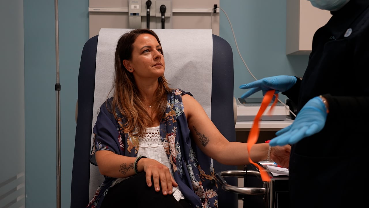 A woman sits with her arm stretched as a masked nurse takes her blood.