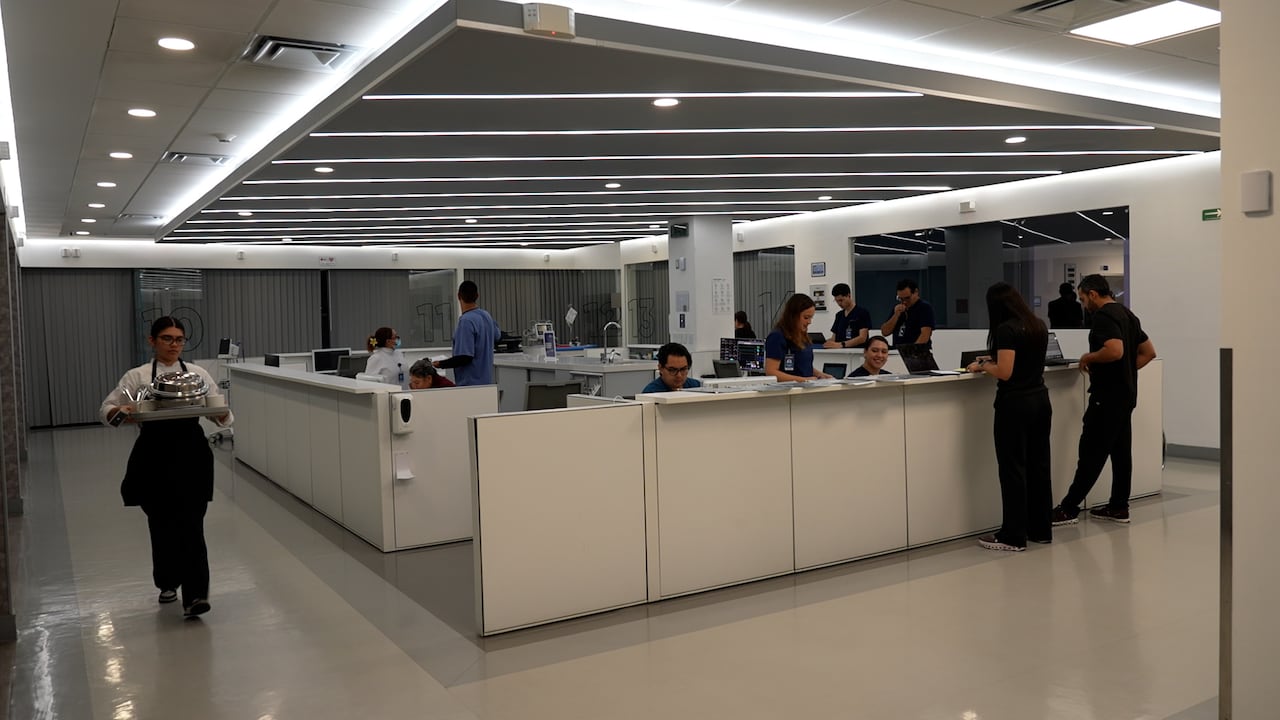 A brithly lit room in a hospital with several curtained-off rooms on the periphery and medical staff desks in the middle.