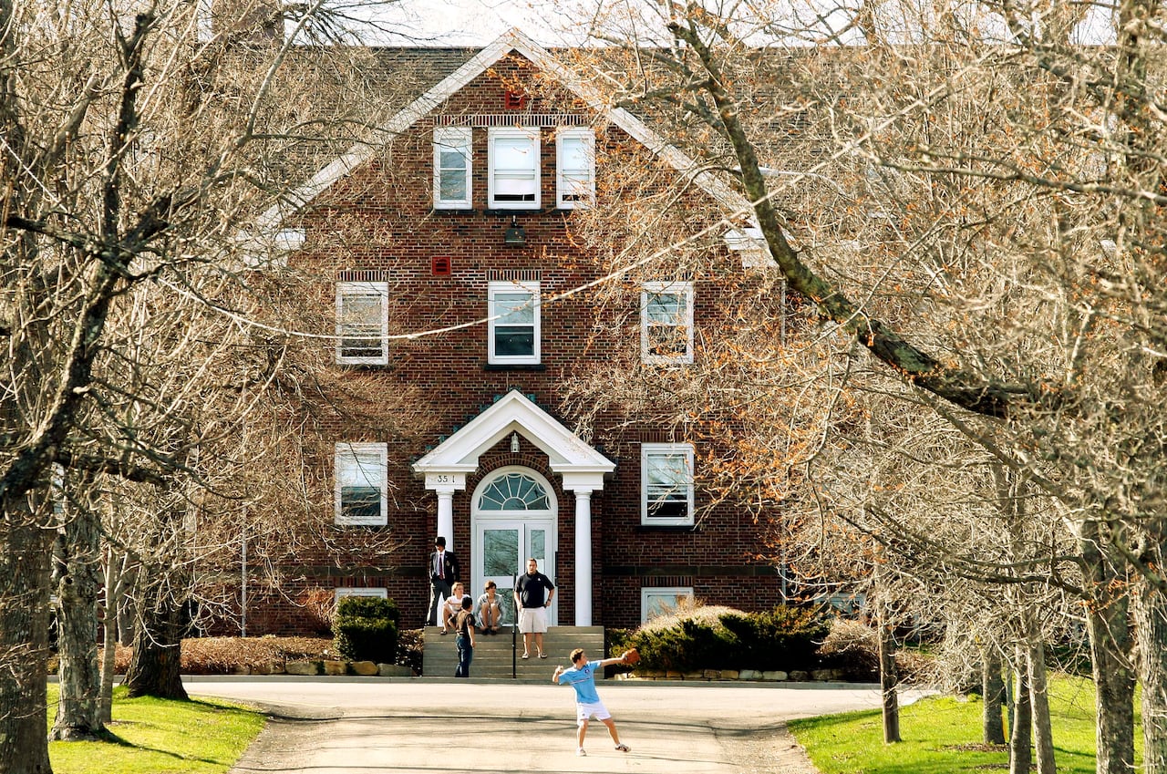 The facade of a brick building is shown through trees. Students stand outside near the front of the building.