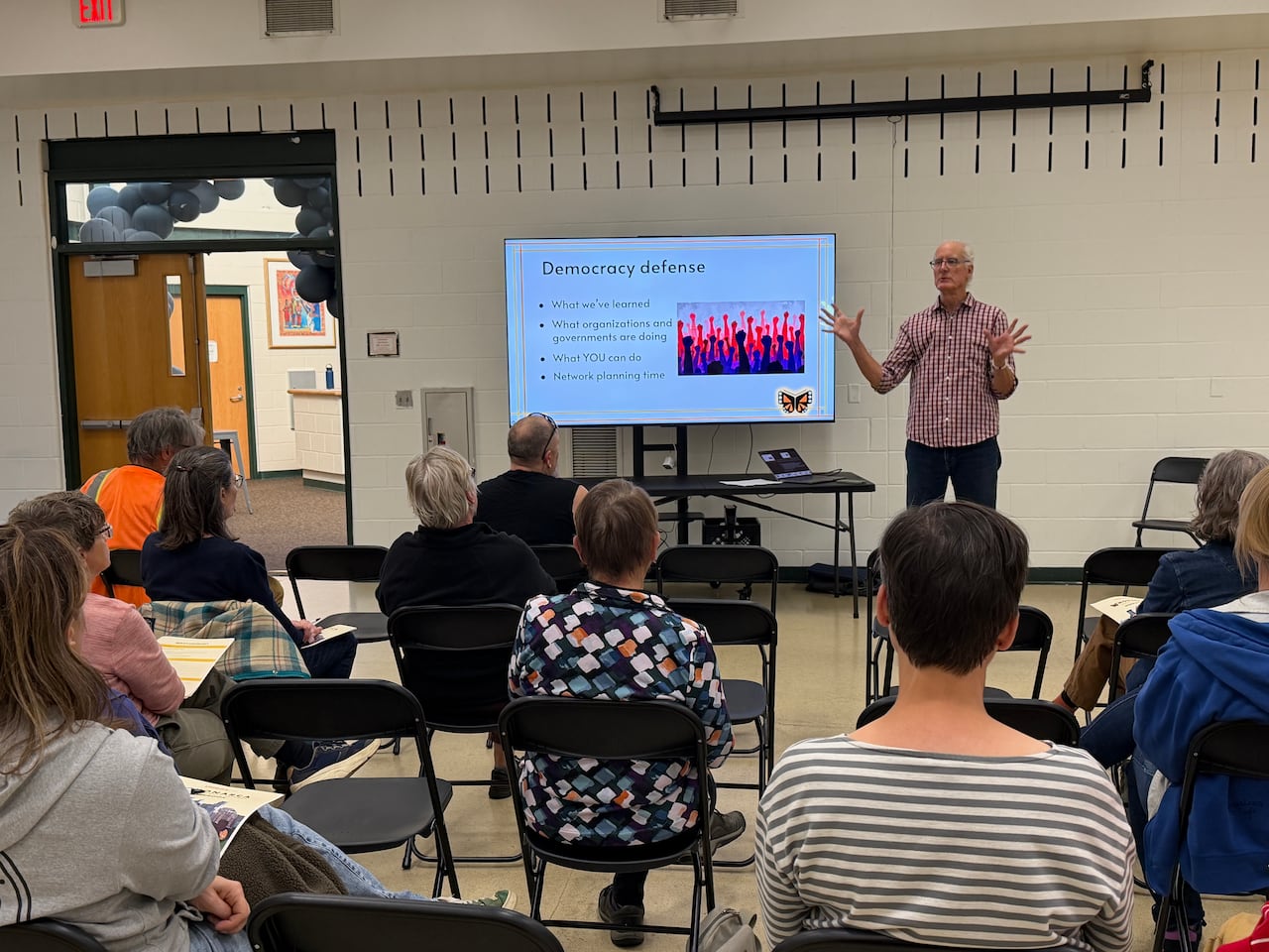 A man stands and talks to a crowd seated in folding chairs in a community space. He is in front of a screen that says 'Democracy defense.'