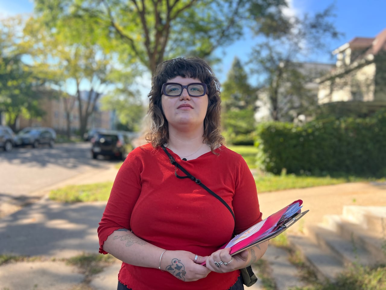 A woman stands on a street with houses and trees, holding a clipboard and looking at the camera.