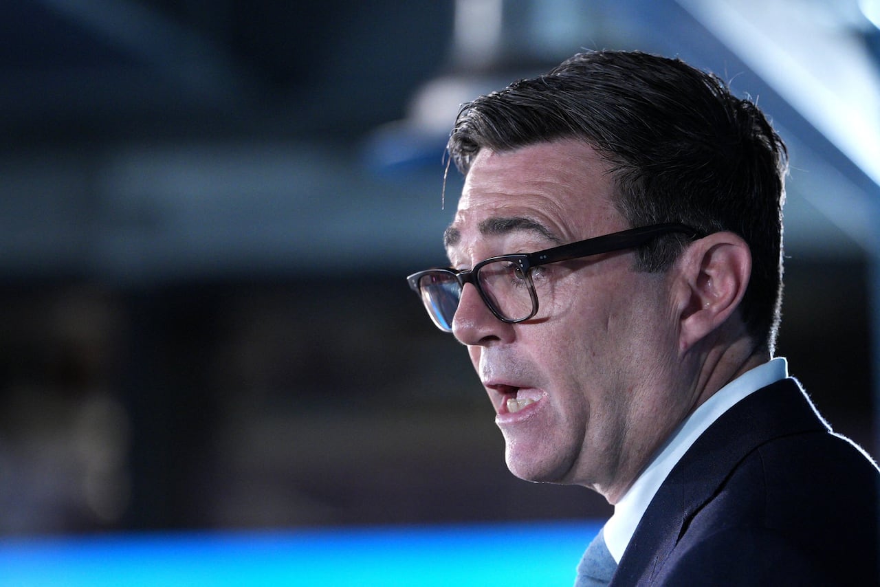 A man with glasses is seen from the side, speaking publicly.