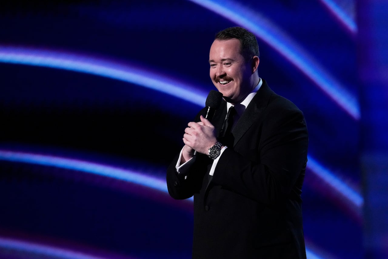 a man in a black tuxedo holds a microphone and smiles on stage as he gives an address