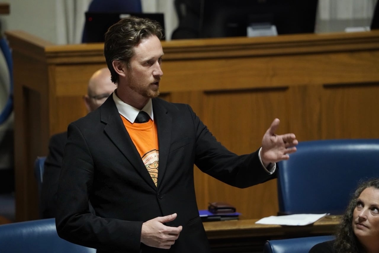 A man in a dark suit and orange shirt stands while speaking during a government legislative session.