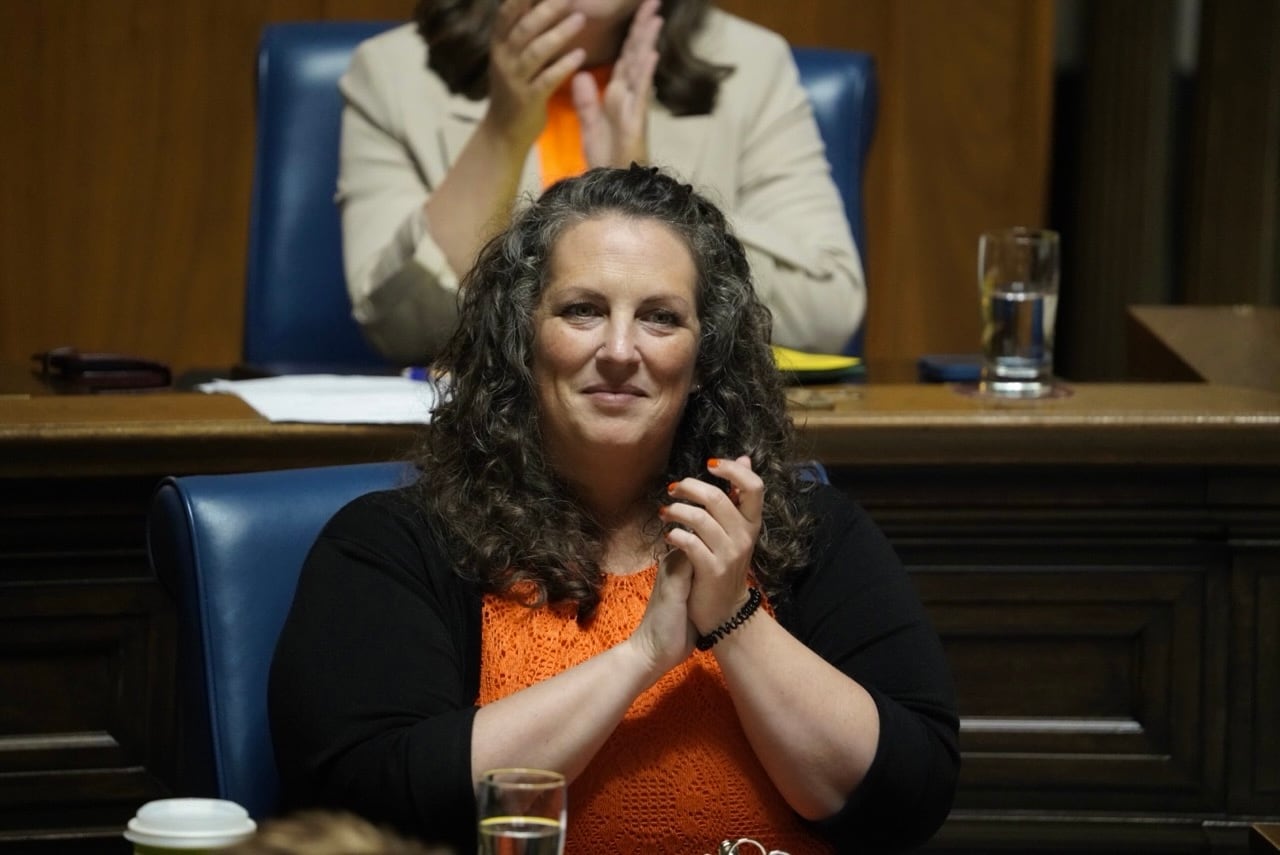 A woman with long curly hair, wearing an orange shirt, claps during a government legislative session.
