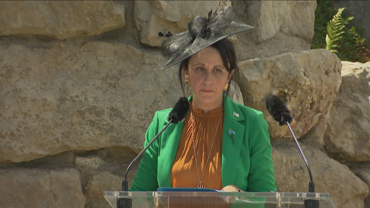 A woman in a suit jacket and black fascinator stands at a lectern with two microphones attached.
