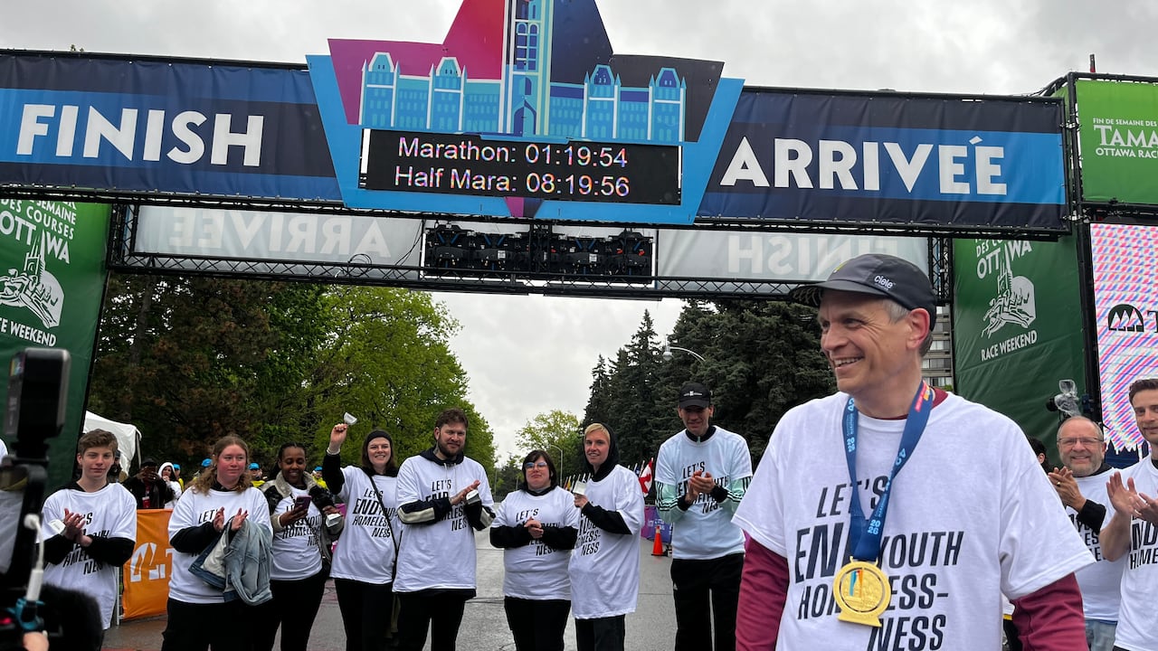 Man with a medal around his next stands close to the camera as a group of others line up in front of a race finish line