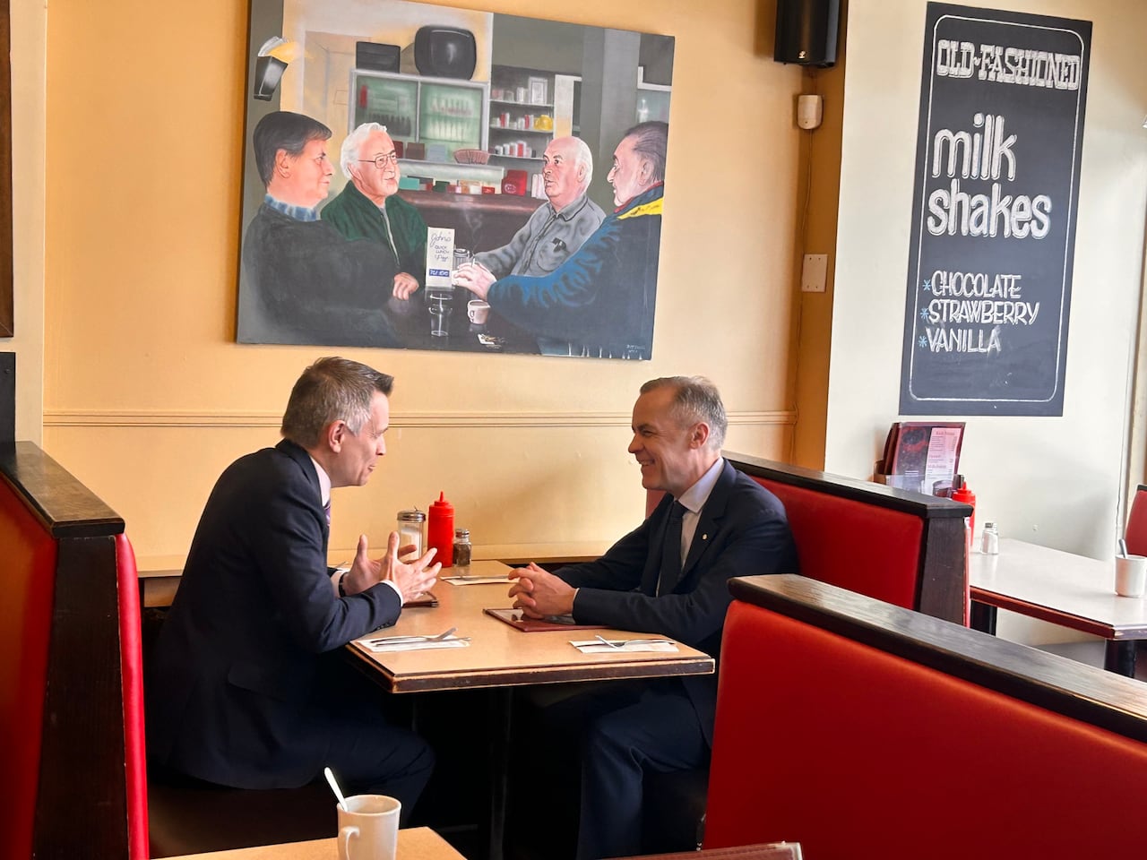 Two men in suits sit facing each other in a diner booth under a painting of four men seated at a diner booth.
