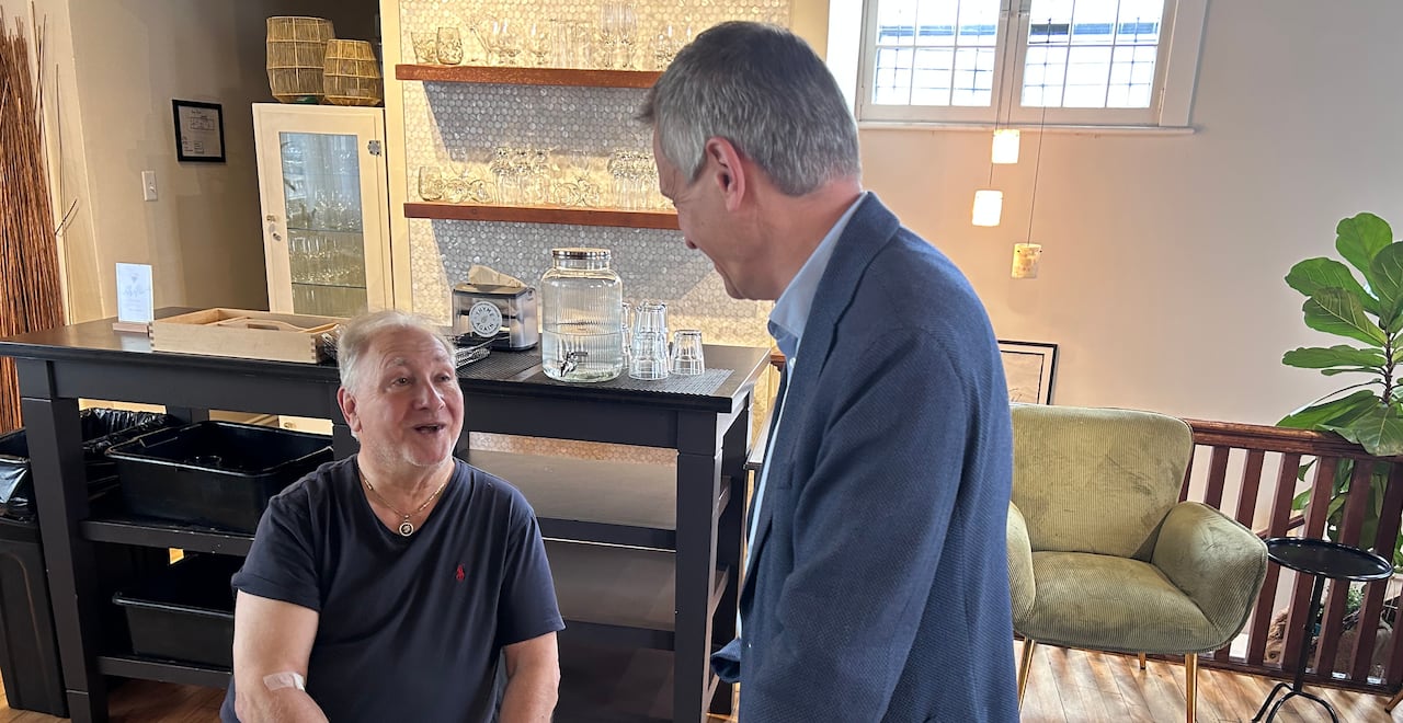 A man stands leans on the back of a chair while talking to a person eating a meal.