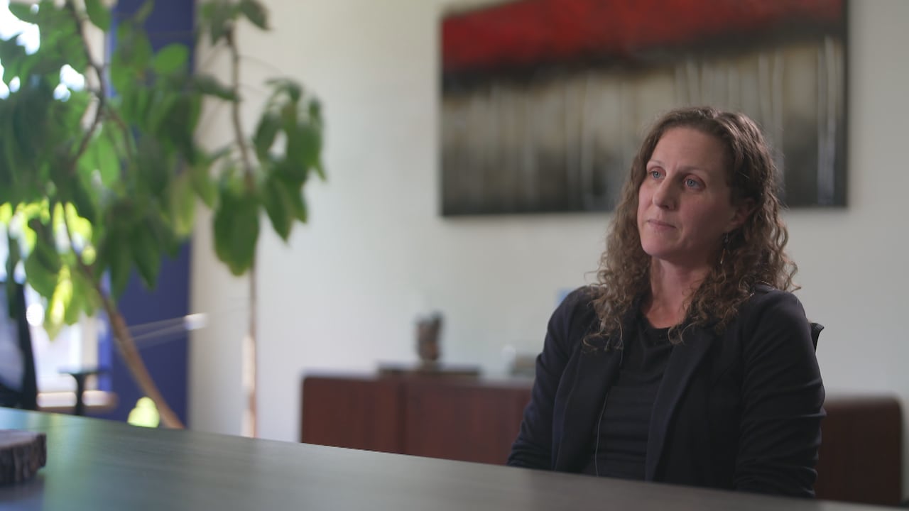 A woman sits at a long, dark, wooden boardroom table with a painting in the background and a tree off to the left. She's wearing a black blazer and has a serious expression.