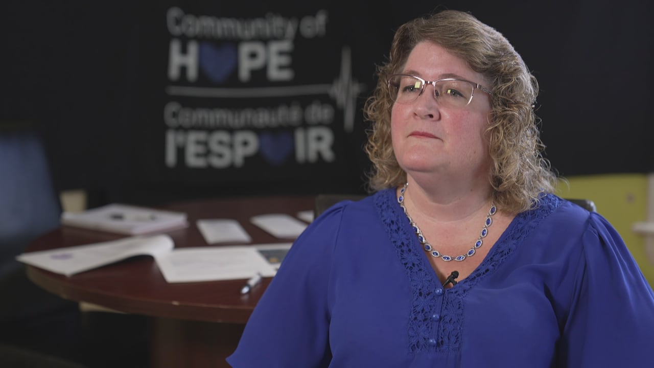 A woman in a blue shirt with blond hair and glasses sits with a "Community of Hope" sign in the background.