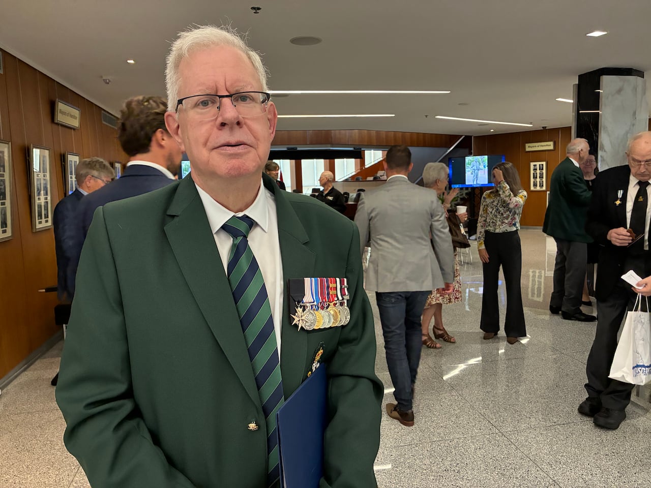 A man wearing a green suit with a number of military medals stands looking at the camera.