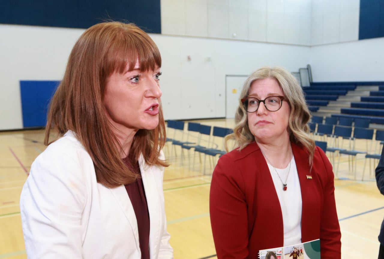A woman in a white blazer standing beside another woman in a red blazer who is holding a document.